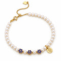 Ilaria white freshwater pearl bracelet with three blue enamel flower beads and a round medallion charm, flat lay on cream